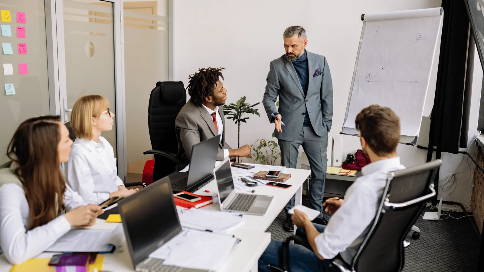 Business team meeting with presenter pointing at colleagues around a laptop-filled table for scheduling