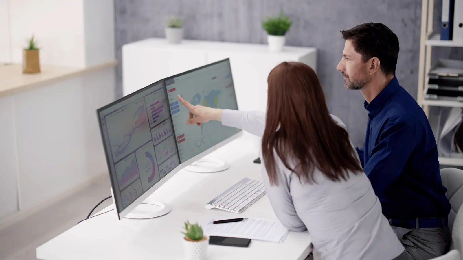 Two colleagues pointing at dual monitors showing charts and maps in office