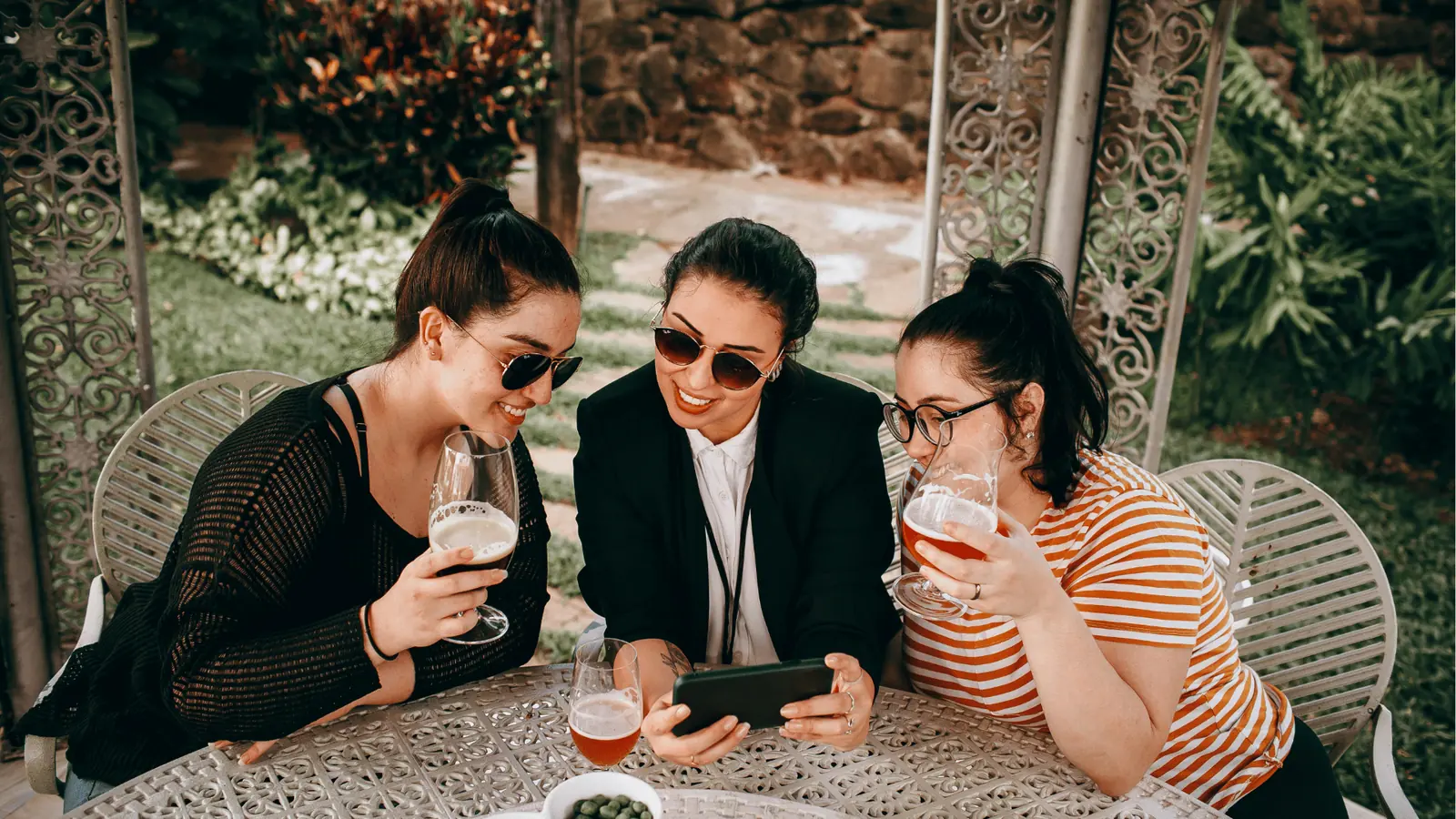 Three friends outdoors at metal table drinking beer and looking at phone