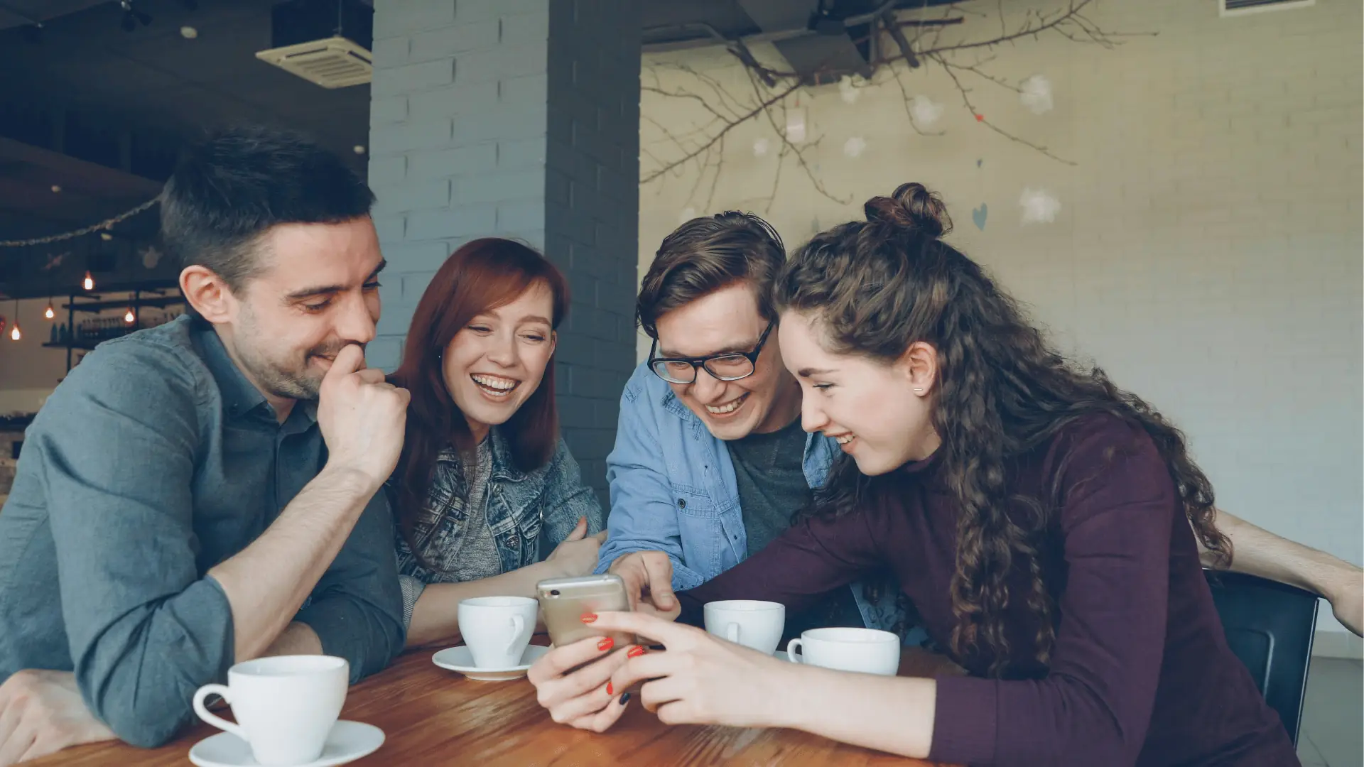 Four friends at a cafe laugh while looking at a smartphone together