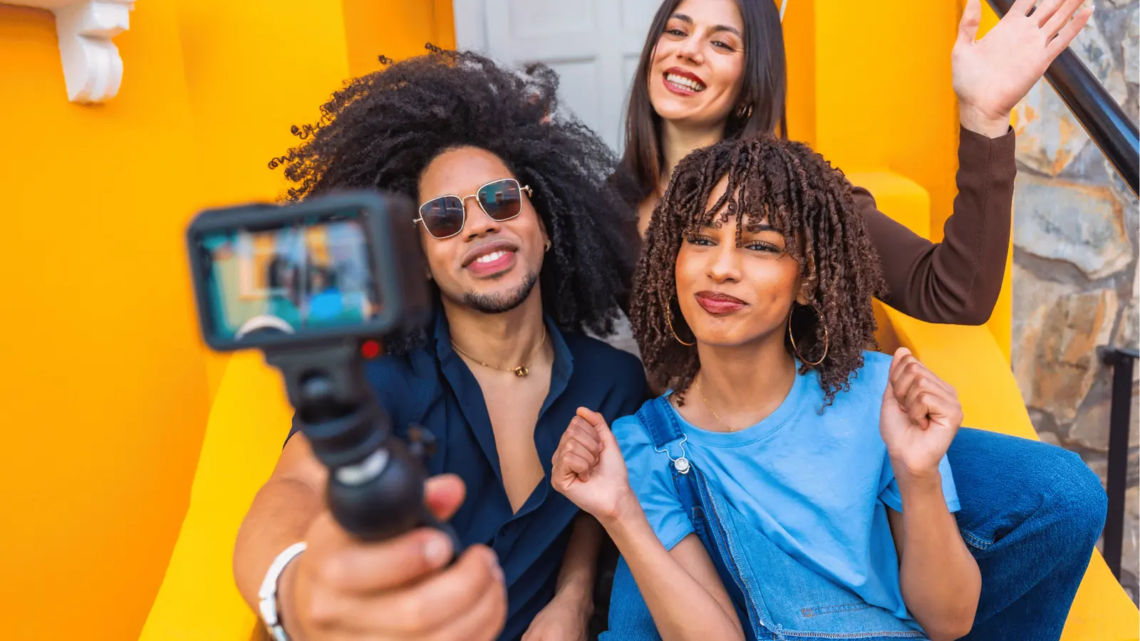 Three friends smiling and recording a vlog together on bright yellow steps