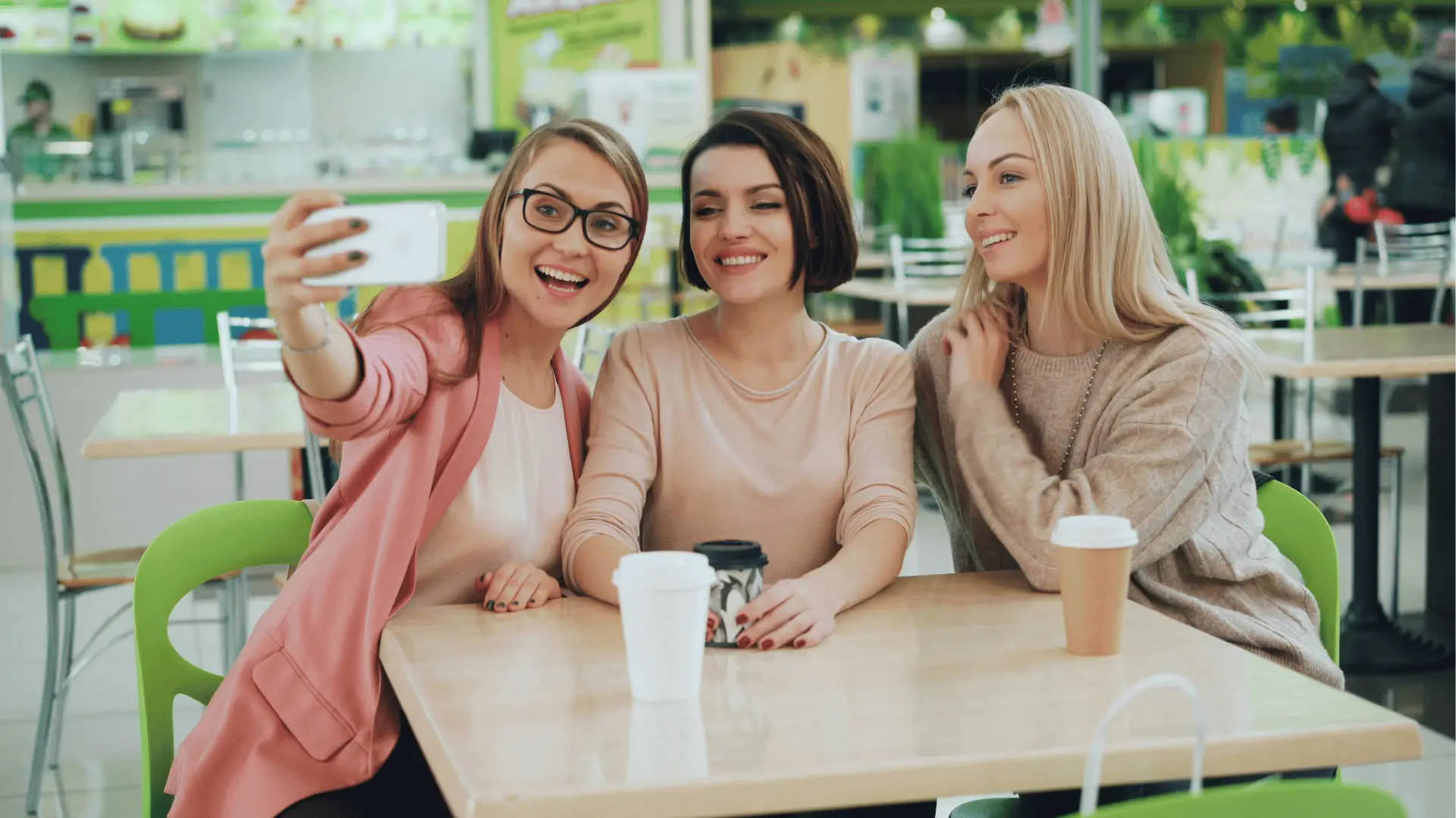 Three young women smiling and taking a selfie together at café table