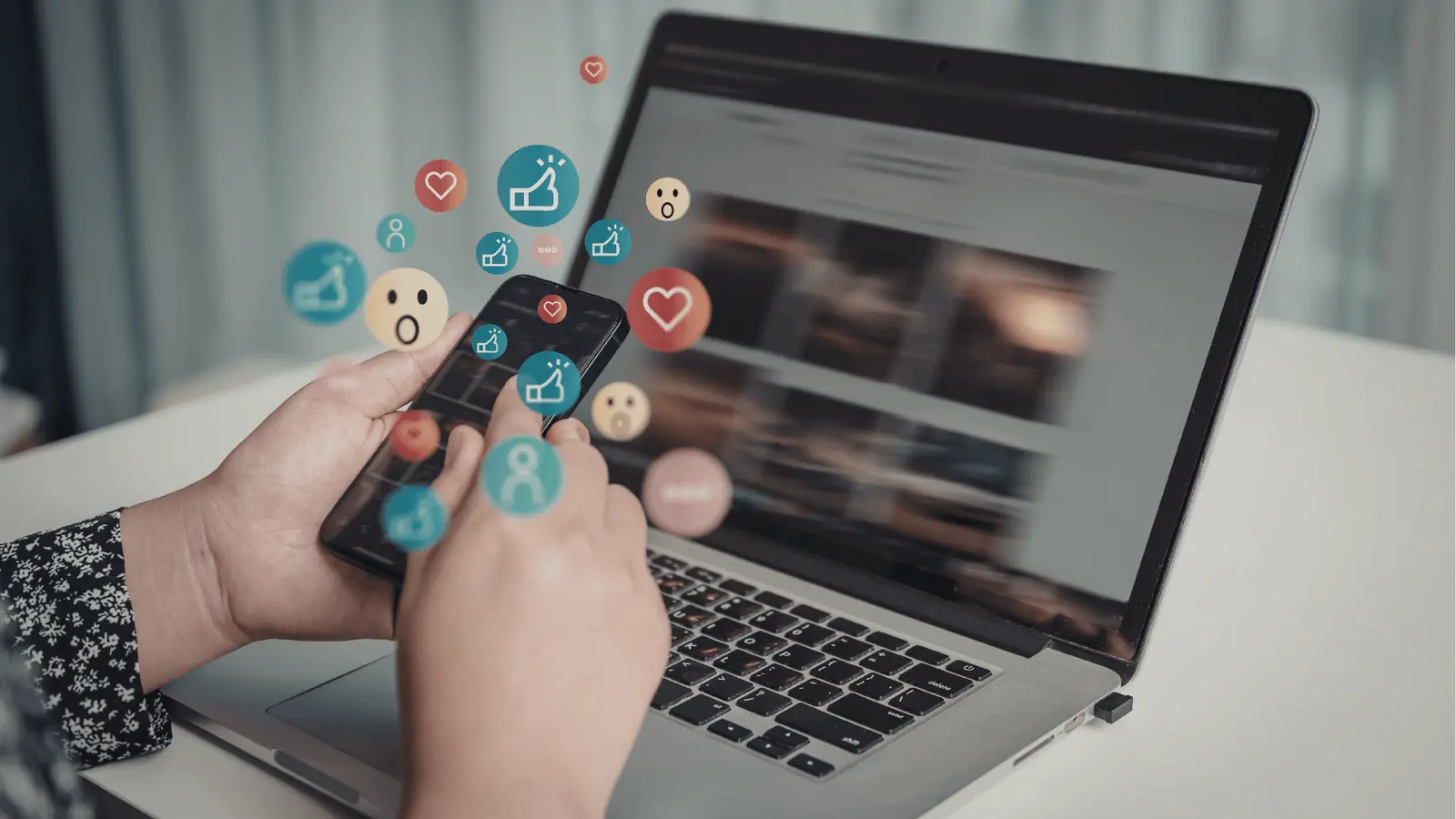 Hands holding smartphone over laptop with floating social reaction icons
