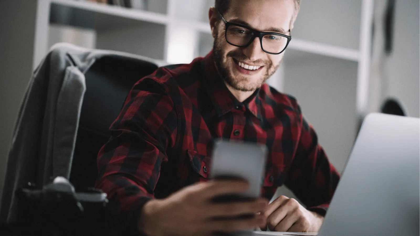 Smiling man in plaid shirt looking at phone beside an open laptop