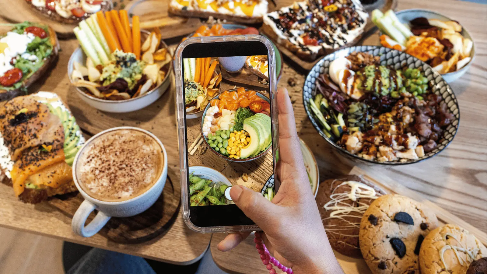 Hand holding smartphone photographing colorful poke bowls, toast, coffee, and cookies