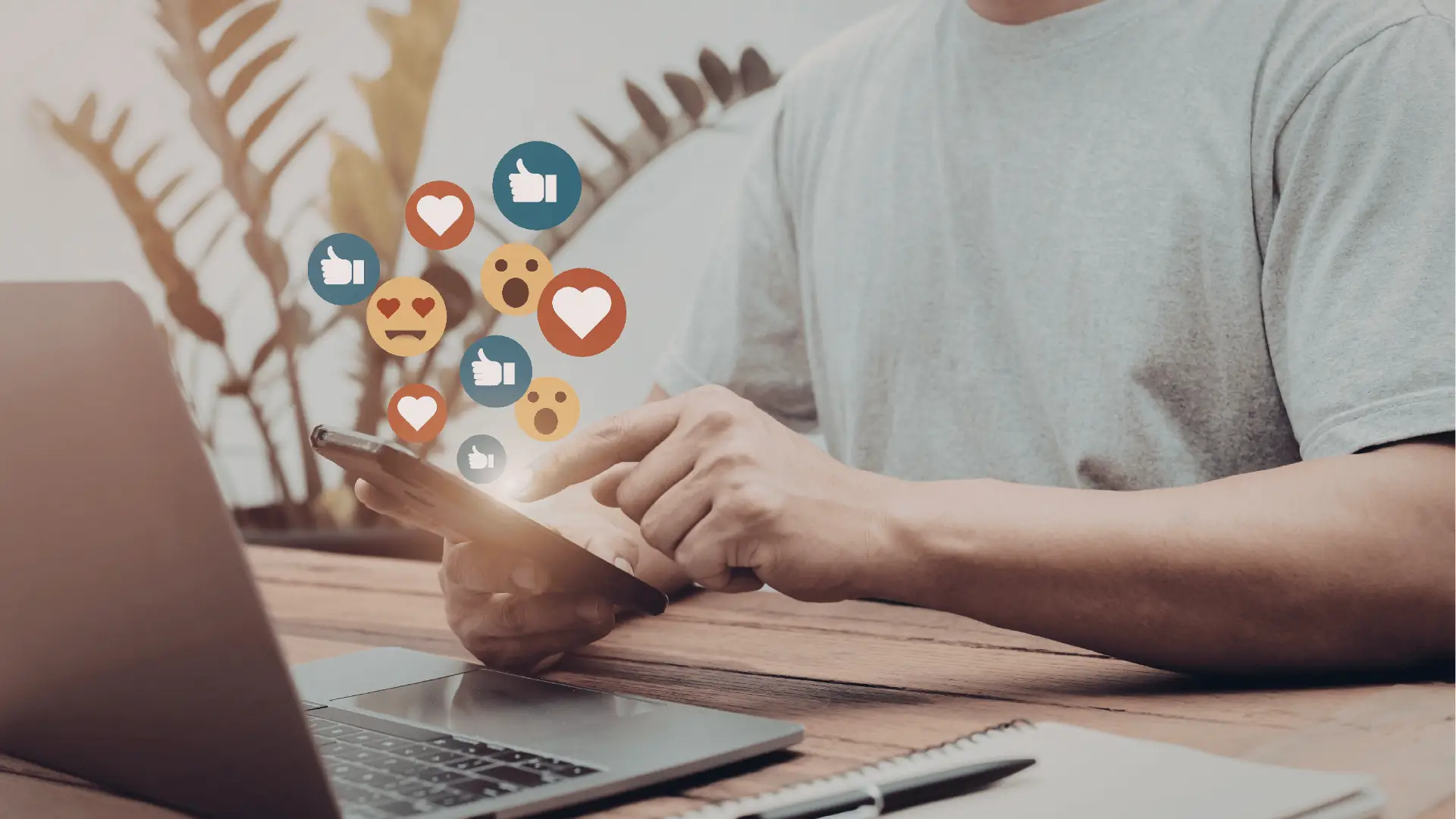 Person tapping smartphone with floating social media reaction icons beside laptop and notebook