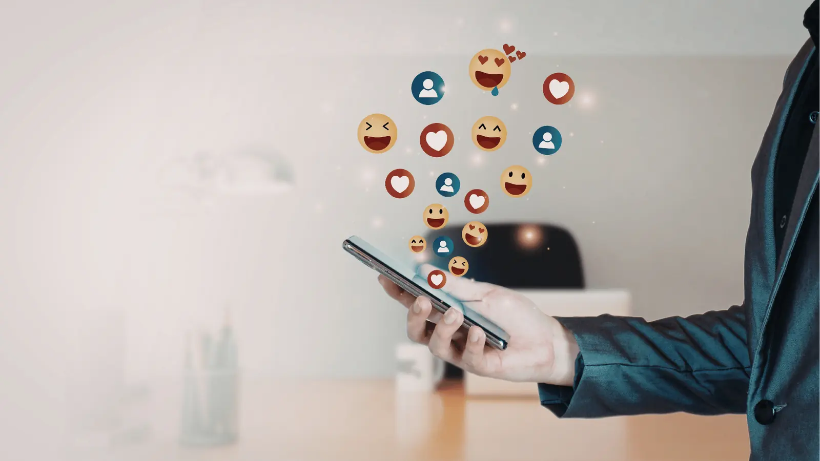 Hand holding smartphone with floating social media reaction icons in blurred office