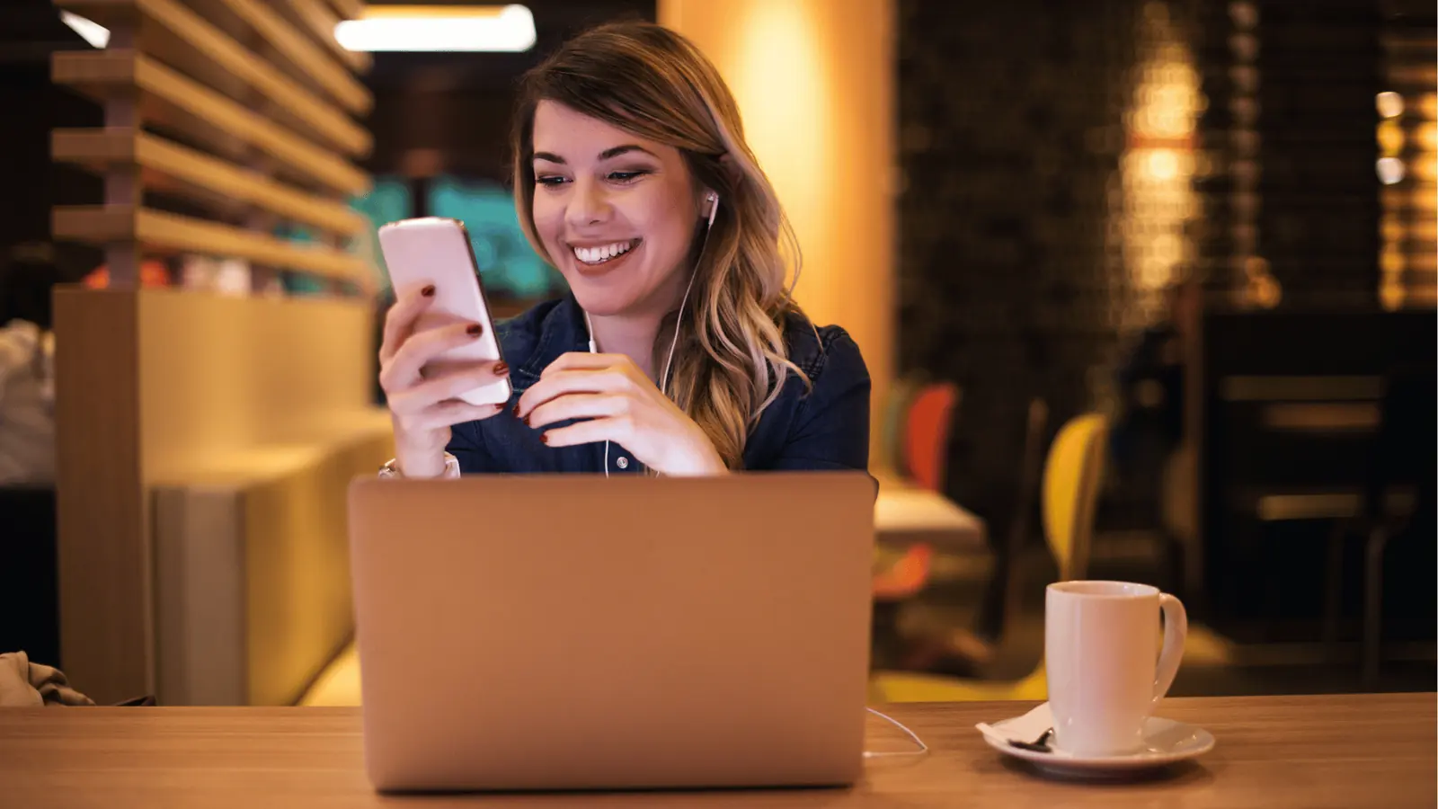 Smiling woman in cafe using smartphone with laptop and coffee on table