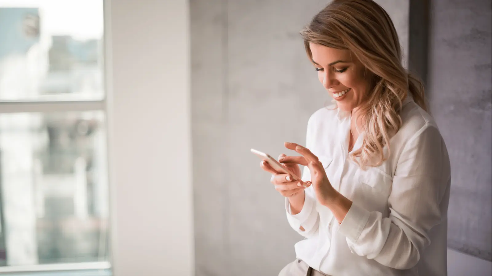Smiling woman in white blouse using a smartphone near a bright window