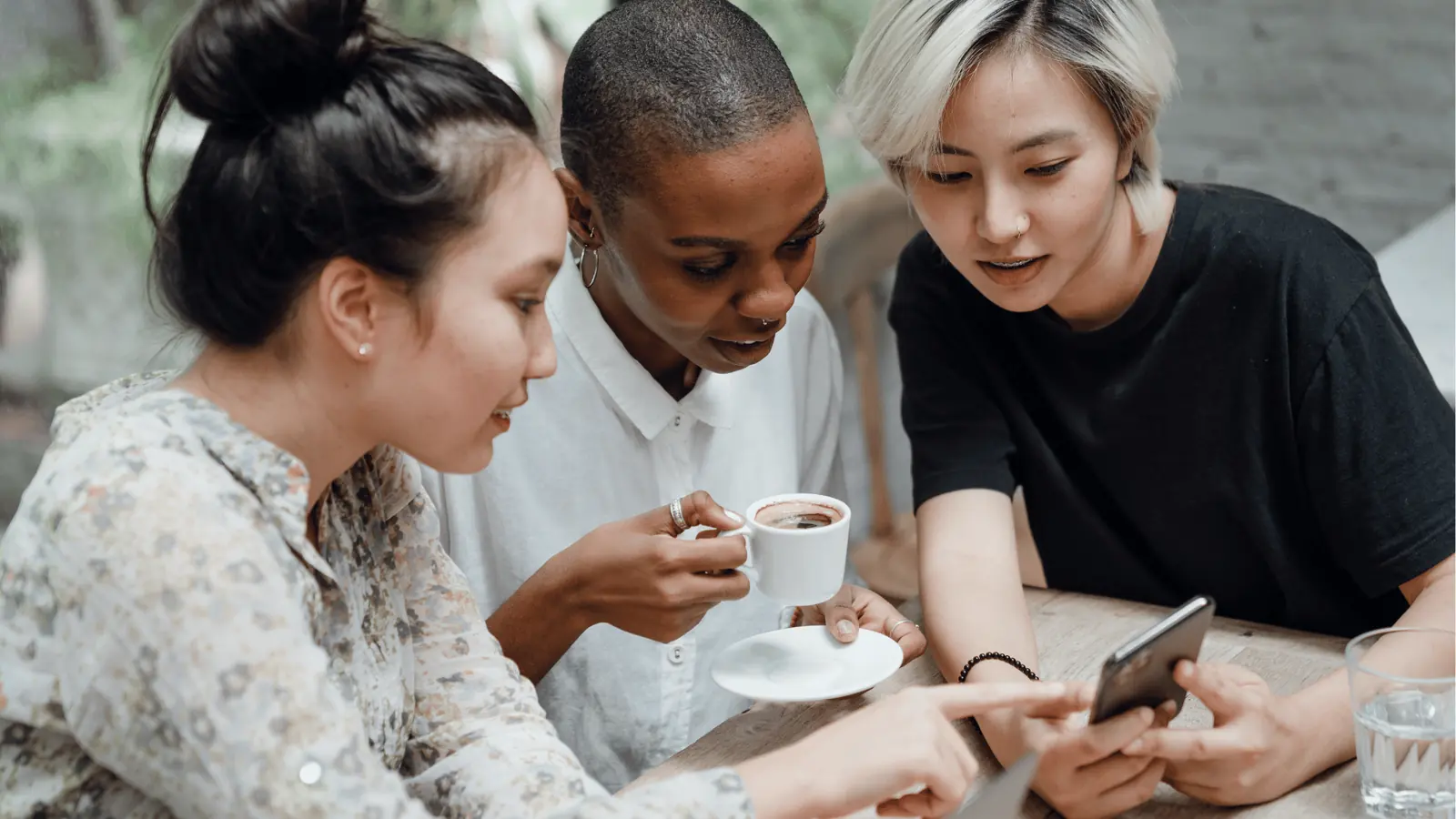 Three friends at a table looking at a smartphone and drinking coffee