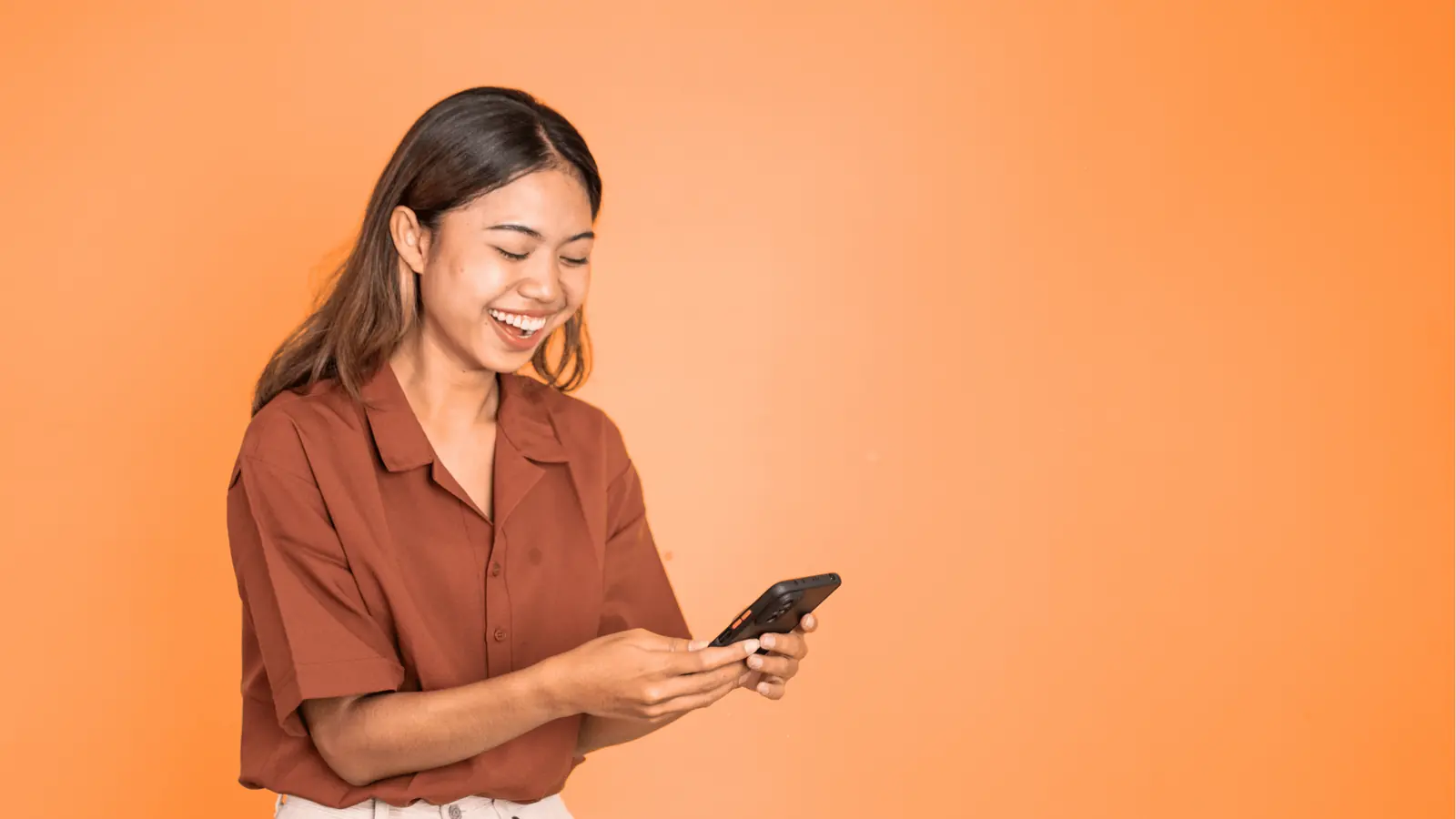 Young woman smiling and looking at smartphone against orange background