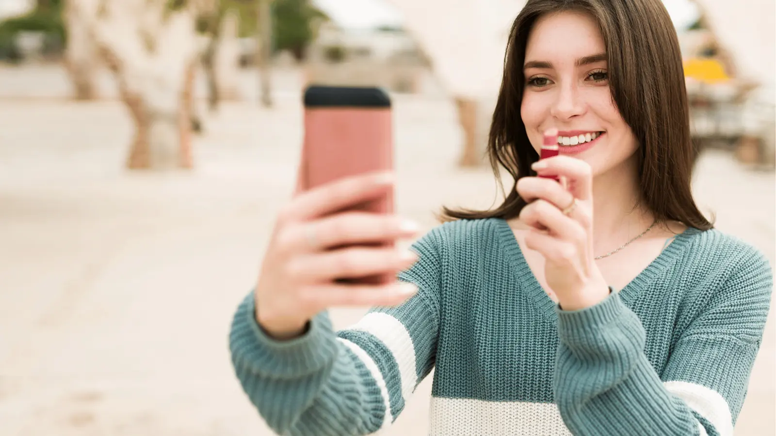 Young woman smiling while taking a lipstick selfie with smartphone outdoors for automation