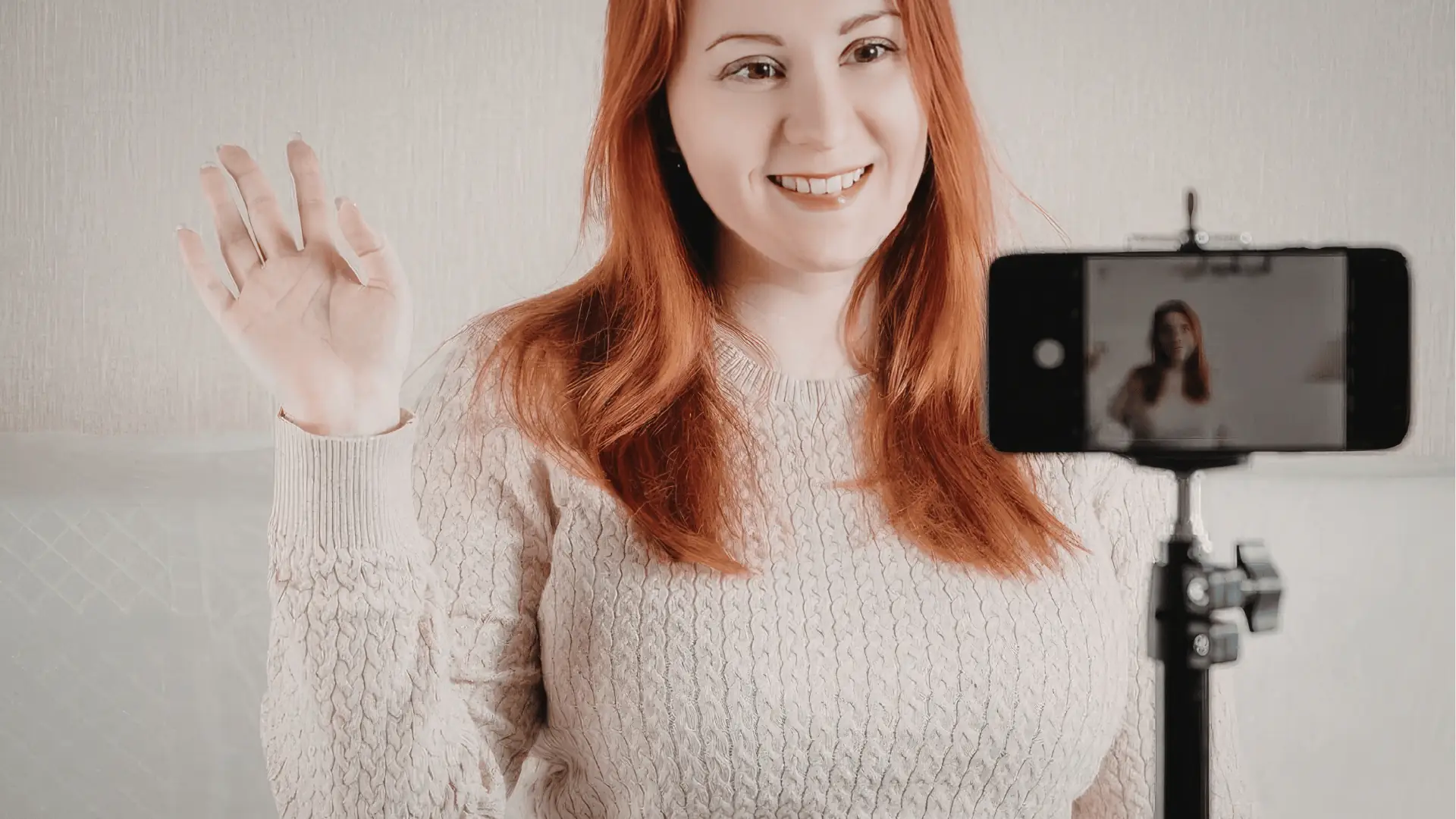 Smiling red-haired woman waving to smartphone camera on tripod