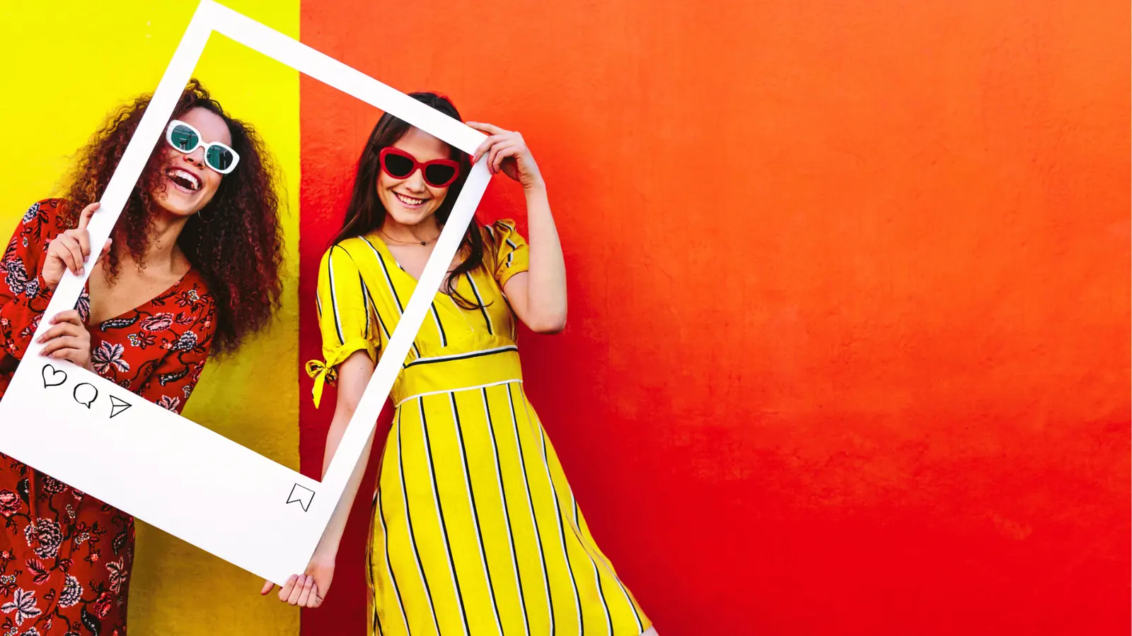 Two smiling women holding a white Instagram-style frame against colorful wall