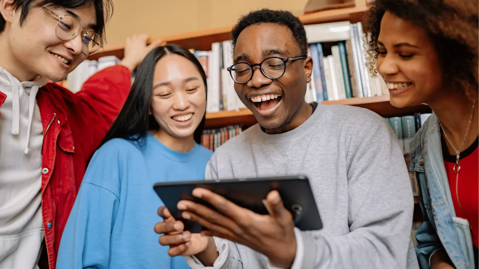 Four young adults smiling together while looking at a tablet in a library