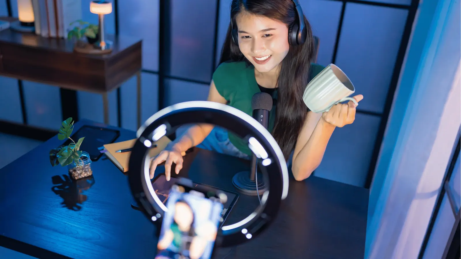 Smiling woman wearing headphones livestreaming at desk with mug and ring light for creative production