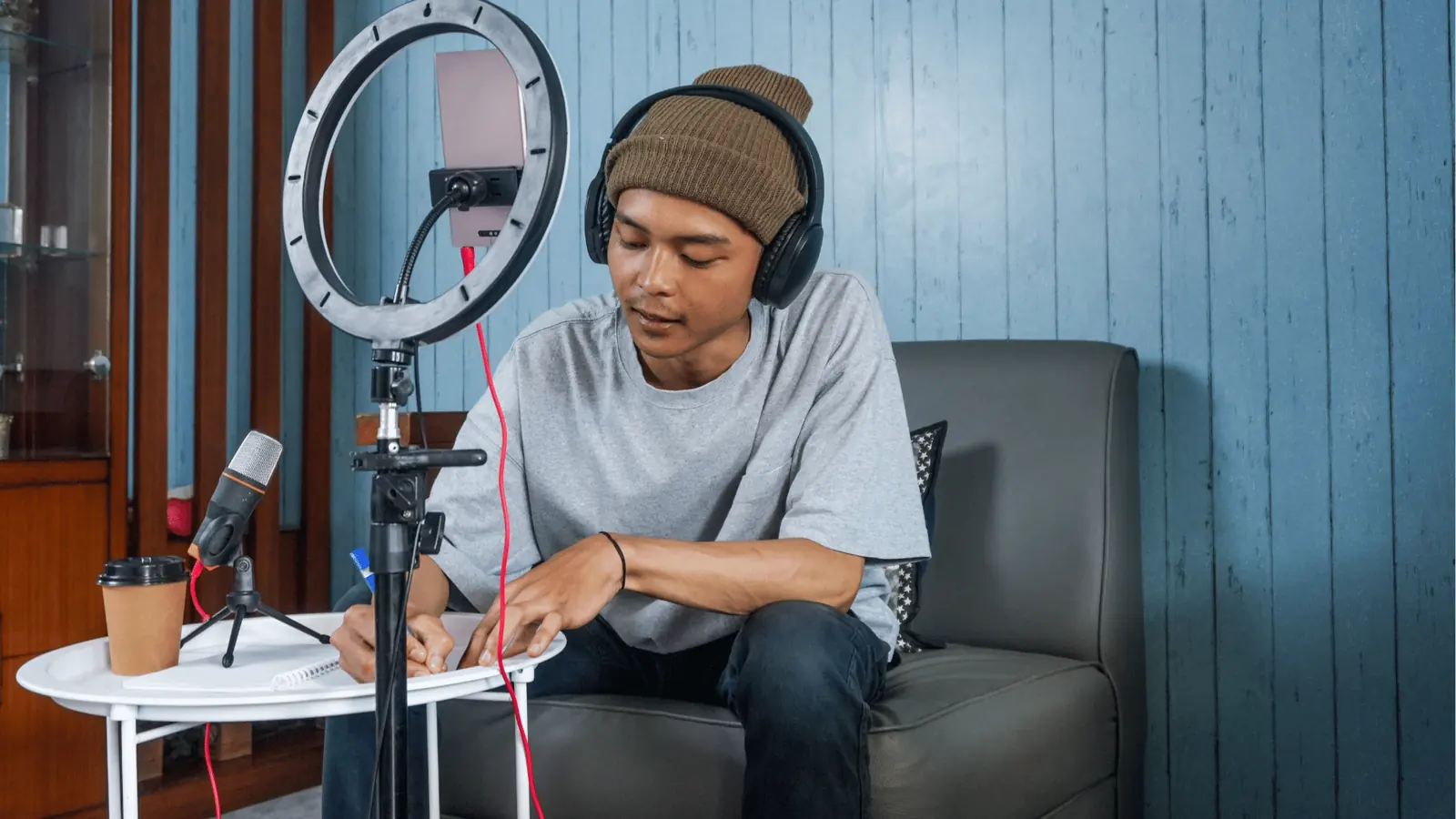 Young man with headphones writing notes while recording with ring light and microphone for asset management