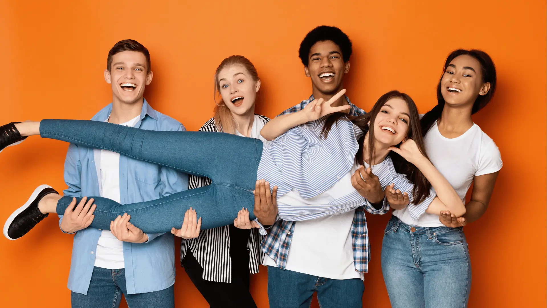 Five young friends smiling while lifting a friend against orange background